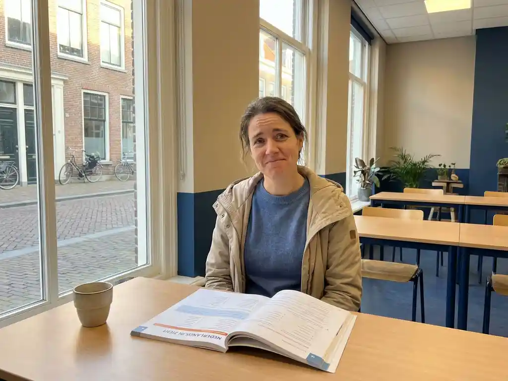 Expat studying Dutch at a bright language school desk with an open textbook and coffee cup, soft afternoon light from a street-facing window.