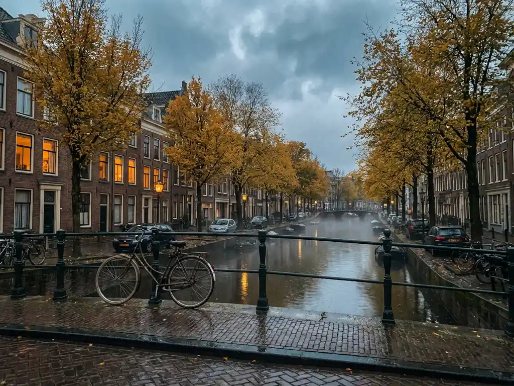 Bicycle leaning on a rain-slicked cobblestone bridge along a misty Dutch canal lined with golden autumn trees and brick rowhouses.