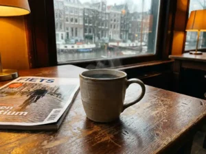 Steaming ceramic mug of dark coffee on a worn wooden café table beside a Dutch cycling magazine, Amsterdam canal blurred in rainy background.
