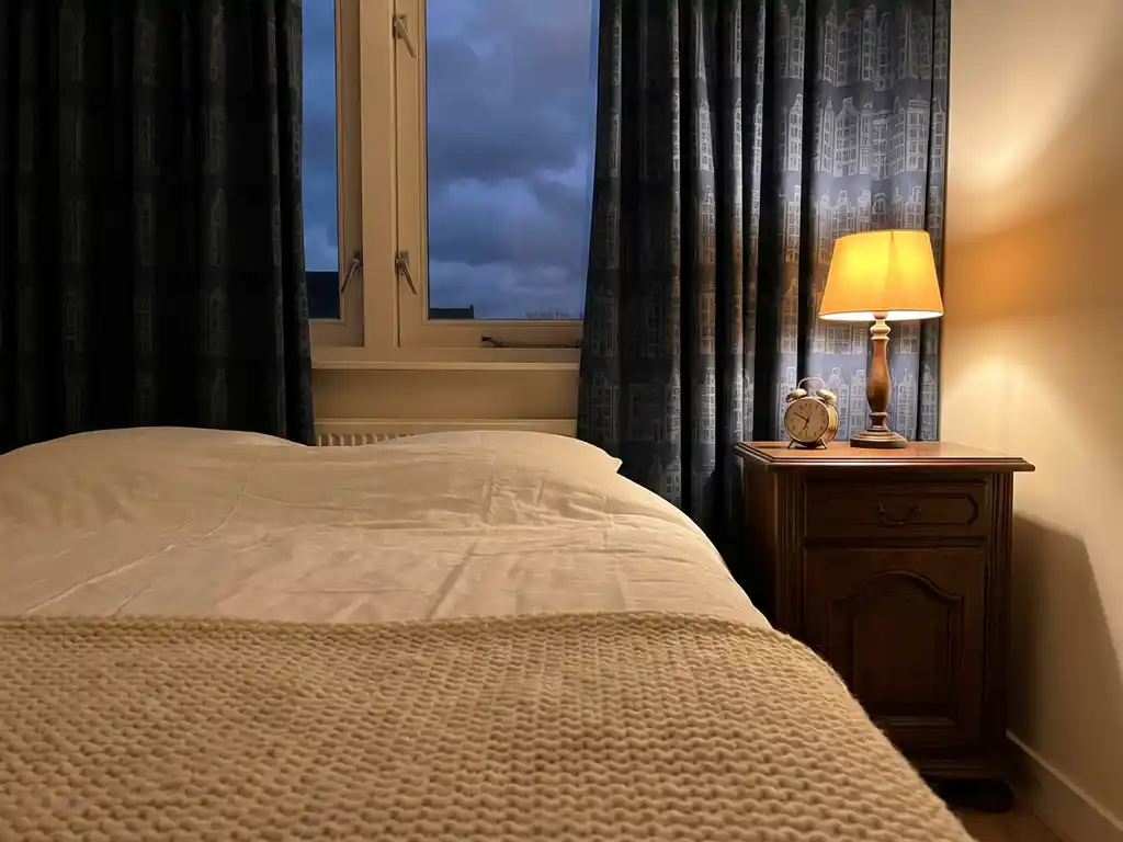 Cozy Dutch bedroom at night with white linens, knitted throw, glowing lamp on wooden nightstand, and canal-house curtains revealing dark evening sky.