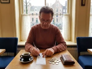 Dutch expat splitting a restaurant bill at an Amsterdam café table with coins, leather wallet, and black coffee beside a canal-view window.